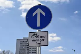 Blaues Verkehrsschild mit weißem Pfeil nach oben und darunter weißes Zusatzschild mit schwarzem Fahrradpiktogramm und Text 'und Anlieger frei' vor bewölktem Himmel und Hochhaus im Hintergrund.