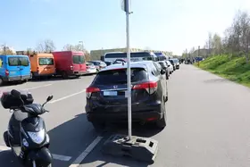 Mehrere Fahrzeuge, darunter ein schwarzer Honda, parken auf einem Gehweg neben einer Straße, ein Verkehrsschild steht auf dem Gehweg, ein Motorroller steht am linken Bildrand.