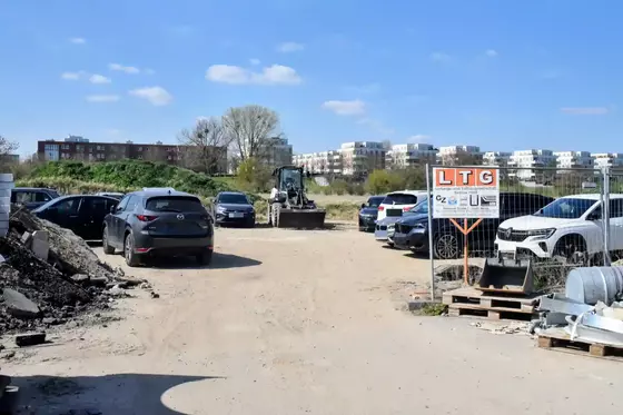 Baustellenbereich mit mehreren geparkten Autos auf unbefestigtem Boden und einem Bagger im Hintergrund