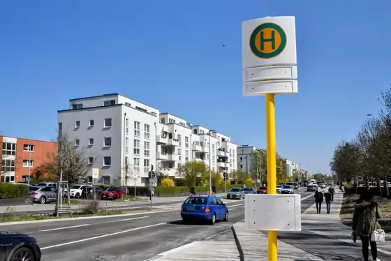 Straße mit mehrstöckigen Wohngebäuden, einem gelb-weißen Haltestellenschild und zwei Fußgängern auf dem Gehweg.