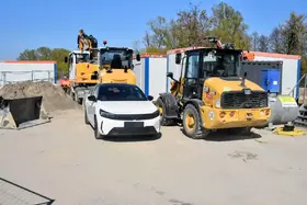 Weißer Pkw steht zwischen zwei gelben Baumaschinen auf einer Baustelle mit Containern im Hintergrund.