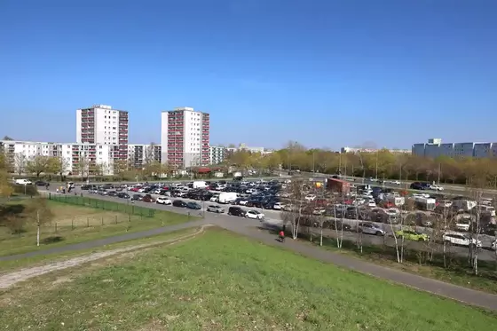 Großer Parkplatz mit vielen geparkten Autos vor mehreren mehrstöckigen Wohngebäuden und einem grasbewachsenen Hügel im Vordergrund
