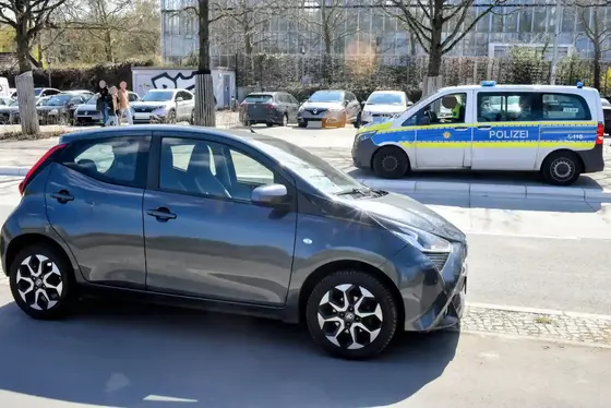 Ein grauer Kleinwagen steht auf einem Gehweg, im Hintergrund steht ein Polizeifahrzeug auf der Straße.