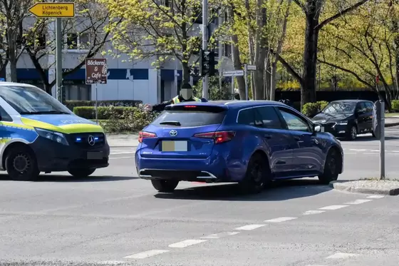 Blauer Toyota fährt an einem Polizeifahrzeug und einer Absperrung vorbei, ein Polizist steht daneben und belehrt den Fahrer.