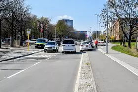 Straße mit mehreren parkenden und fahrenden Autos sowie einem Radfahrer auf dem Radweg bei klarem Himmel.