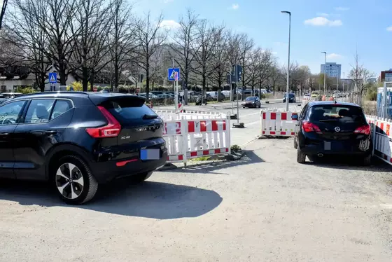 Zwei schwarze Autos parken auf einem unbefestigten Platz neben einer abgesperrten Baustelle mit weißen und roten Absperrungen und Verkehrsschildern im Hintergrund.
