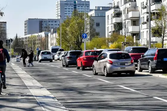 Mehrere Autos fahren in einer zweispurigen Straße an einem Wohngebiet mit mehrstöckigen Gebäuden und Bäumen.