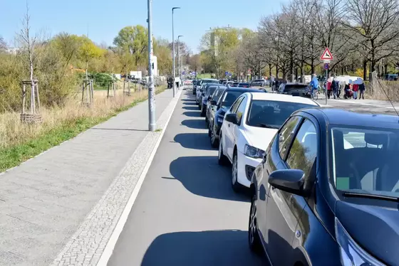 Mehrere Autos parken auf einem Radweg neben einem Gehweg und einer Straße mit Bäumen und Fußgängern im Hintergrund.