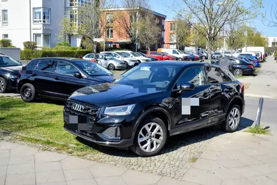 Zwei schwarze Autos, eines auf dem Gehweg und eines auf dem angrenzenden Grünstreifen geparkt, umgeben von weiteren parkenden Fahrzeugen und Wohngebäuden im Hintergrund.
