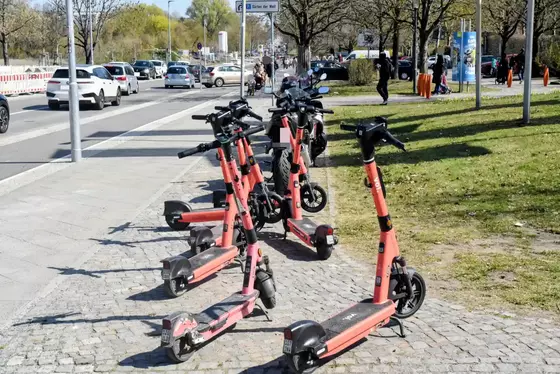Mehrere orangefarbene E-Scooter stehen auf einem gepflasterten Gehweg neben einer Straße mit parkenden Autos und Bäumen im Hintergrund.