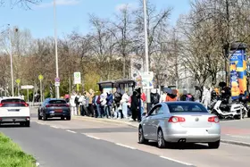 Lange Schlange von Menschen an einer Bushaltestelle neben einer Straße mit mehreren fahrenden Autos und Bäumen im Hintergrund.