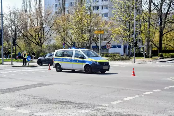 Polizeiwagen steht auf einer Kreuzung mit zwei orangefarbenen Verkehrskegeln im Vordergrund und Bäumen sowie Gebäuden im Hintergrund.