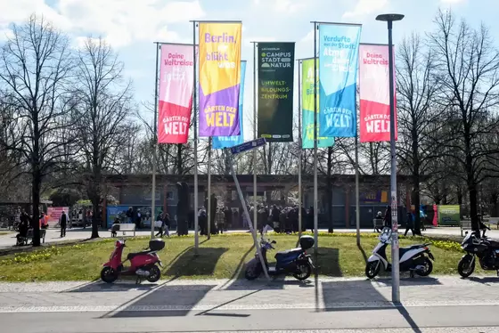 Mehrere bunte Fahnen mit Aufschriften zu den Gärten der Welt stehen auf einer Wiese vor einem Gebäude mit Menschen und geparkten Rollern im Vordergrund.