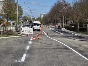 Straße mit Baustellenabsperrung, mehreren orange-weißen Verkehrskegeln und einem weißen Transporter, daneben zwei Personen in orangefarbener Arbeitskleidung.