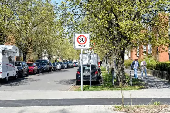 Straße mit parkenden Autos und Bäumen, im Vordergrund ein Verkehrsschild mit der Aufschrift '30 ZONE', dahinter ein weiteres Schild mit einem Durchfahrtsverbot verdeckt.