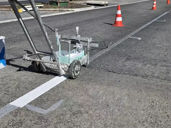 Markierungsgerät auf Asphalt mit frisch gezogener weißer Linie und mehreren orange-weißen Verkehrskegeln im Hintergrund.