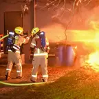 Drei Feuerwehrleute in Schutzkleidung und Helmen löschen mit einem Schlauch ein Feuer im Freien.