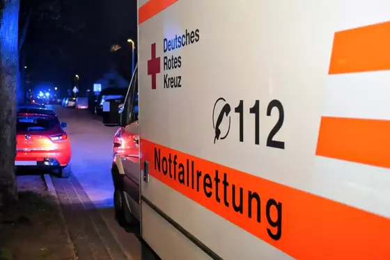 Seitenansicht eines Rettungswagens mit der Aufschrift 'Deutsches Rotes Kreuz', '112' und 'Notfallrettung' bei Nacht auf einer Straße mit parkenden Autos und Blaulicht im Hintergrund.