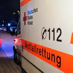 Seitenansicht eines Rettungswagens mit der Aufschrift 'Deutsches Rotes Kreuz', '112' und 'Notfallrettung' bei Nacht auf einer Straße mit parkenden Autos und Blaulicht im Hintergrund.