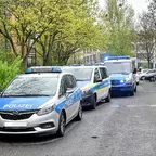 Drei hintereinander geparkte Polizeifahrzeuge auf einer Straße neben einem Gehweg mit Bäumen und parkenden Autos