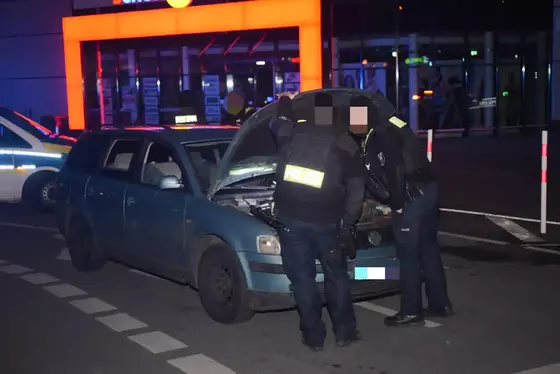 Zwei Polizisten durchsuchen bei Nacht den geöffneten Motorraum eines grünen VW-Kombis auf einer Straße vor einem beleuchteten Gebäude