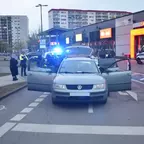 VW mit geöffneten Türen steht auf einer Straße vor einem Einkaufszentrum, mehrere Polizisten und ein Polizeifahrzeug mit Blaulicht sind anwesend.