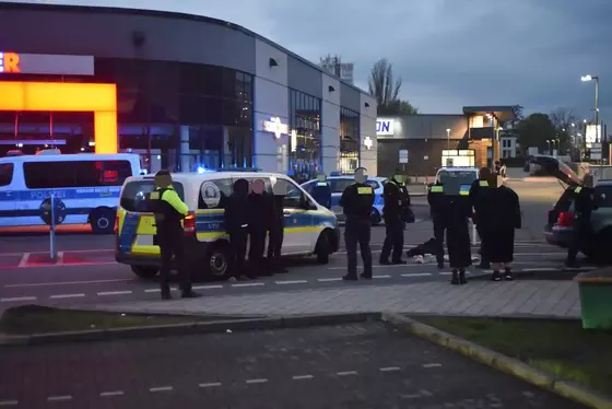 Drei festgenommene Personen stehen neben einem Polizeifahrzeug, umgeben von mehreren Polizeibeamten auf einem Parkplatz vor einem Gebäude bei Dämmerung.