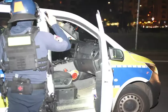 Polizist mit Schutzweste und Helm steht neben offenem Fahrertür eines Polizeifahrzeugs mit Glassplittern im Innenraum bei Nacht