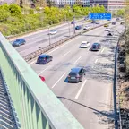 Blick von einer Brücke auf eine mehrspurige Autobahn mit mehreren Fahrzeugen und einem blauen Verkehrsschild im Hintergrund.