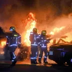 Feuerwehrleute in Schutzkleidung löschen brennende Fahrzeuge bei Nacht.