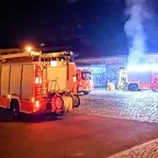 Mehrere Feuerwehrfahrzeuge stehen nachts auf einem gepflasterten Platz vor einem Gebäude, hinter einem Fahrzeug steigt Rauch auf, vermutlich von einem brennenden Müllcontainer.