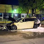 Ausgebrannter weißer BMW-Kombi mit geöffnetem Motorhaubenbereich auf nasser Straße bei Nacht, im Hintergrund ein Zaun und Bäume.