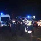 Polizisten und Rettungskräfte mit Warnwesten stehen nachts auf einer Straße vor Einsatzfahrzeugen mit Blaulicht.