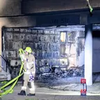 Durch Brand beschädigte Fassade eines Gebäudes mit sichtbaren Rußspuren und einem Feuerwehrmann in Schutzkleidung, der eine Schlauchleitung aufrollt.