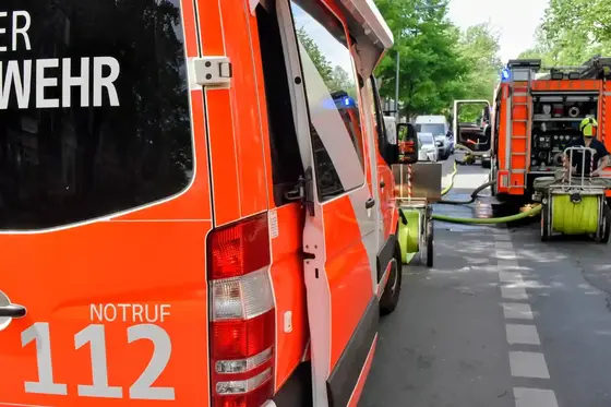 Rückansicht eines Feuerwehrfahrzeugs mit geöffneter Tür und angeschlossenen Schläuchen auf einer Straße mit weiteren Feuerwehrfahrzeugen und Einsatzkräften im Hintergrund.