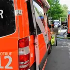 Rückansicht eines Feuerwehrfahrzeugs mit geöffneter Tür und angeschlossenen Schläuchen auf einer Straße mit weiteren Feuerwehrfahrzeugen und Einsatzkräften im Hintergrund.