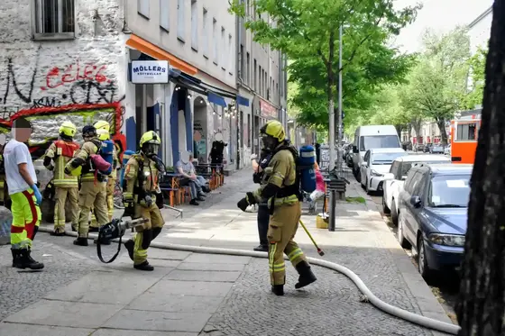 Feuerwehrleute in Schutzkleidung mit Atemschutzgeräten auf einem Bürgersteig vor einem Gebäude in einer Stadtstraße mit geparkten Autos.