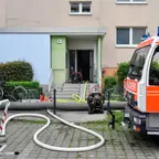 Feuerwehrauto vor einem Wohnhaus mit mehreren Schläuchen, die in den Eingang führen, und Fahrrädern neben dem Hauseingang.