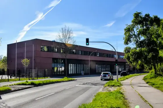 Ampelanlage mit grünem Licht vor einem modernen, zweigeschossigen Gebäude neben einer Straße mit zwei Autos und einem Gehweg mit Bäumen.