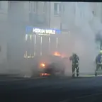 PKW steht in Flammen vor einem Gebäude mit dem Schild 'HOOKAH WORLD', zwei Feuerwehrleute löschen den Brand mit Atemschutzgeräten und Löschschläuchen.