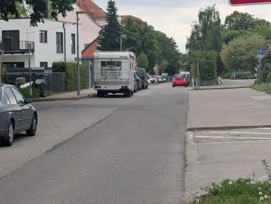 Straße mit geparkten Autos und einem Wohnmobil vor Wohnhäusern und Bäumen.
