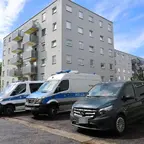 Mehrere Polizeifahrzeuge, darunter ein Kriminaltechnik-Wagen, stehen vor einem mehrstöckigen Wohngebäude mit Balkonen unter blauem Himmel.