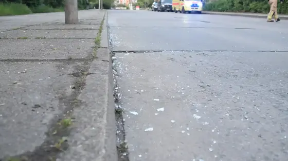 Nahaufnahme von Glassplittern auf einer Straße neben einem Gehweg mit Bäumen und einem Rettungswagen im Hintergrund