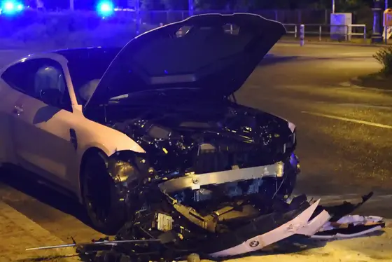 Ein weißes Auto mit geöffneter Motorhaube und stark beschädigter Frontpartie steht nachts auf der Straße, im Hintergrund sind Blaulichter zu sehen.
