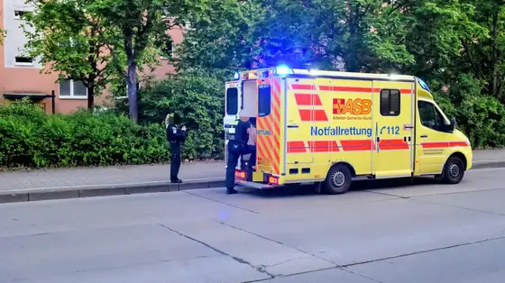 Polizisten neben einem gelben Rettungswagen mit Blaulicht auf einer Straße vor einem Gehweg mit Bäumen und einem Wohngebäude im Hintergrund