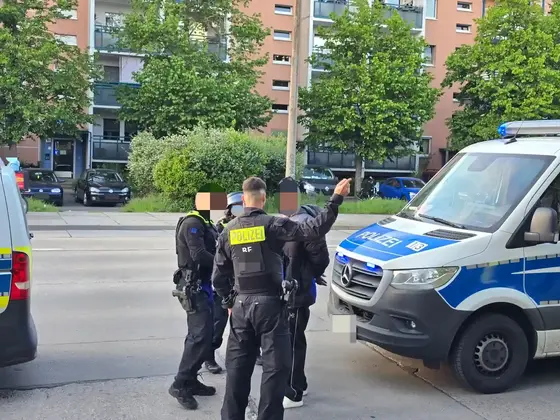 Drei Polizisten in schwarzer Einsatzkleidung führen eine Festnahme auf einem Bürgersteig vor einem Wohnhaus durch, ein Festgenommener mit erhobenem Arm steht vor einem Polizeifahrzeug.