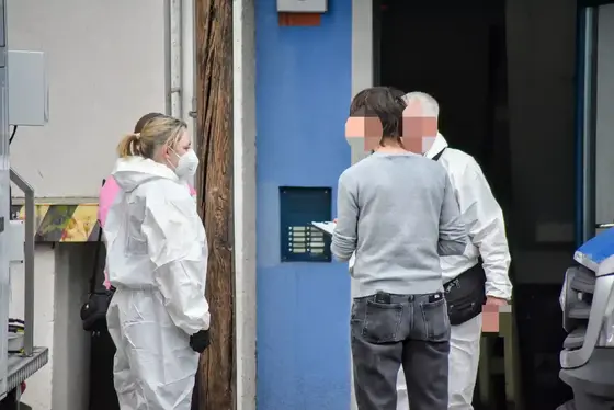 Vier Personen, darunter zwei in weißen Schutzanzügen, stehen vor einem blauen Hauseingang und unterhalten sich.