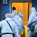 Vier Personen in weißen Schutzanzügen betreten ein Gebäude mit der Nummer 5, rechts daneben steht ein Polizeifahrzeug.
