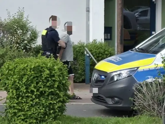 Polizist führt mutmaßlich Tatverdächtigen in kurzen Hosen und T-Shirt an einem Wohnhaus vorbei zu einem Polizeifahrzeug.