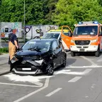 Schwarzer Toyota mit Unfallschaden an der Front steht auf der Straße neben einem Feuerwehrfahrzeug mit geöffneter Tür und einer Person in Feuerwehr-Kleidung.